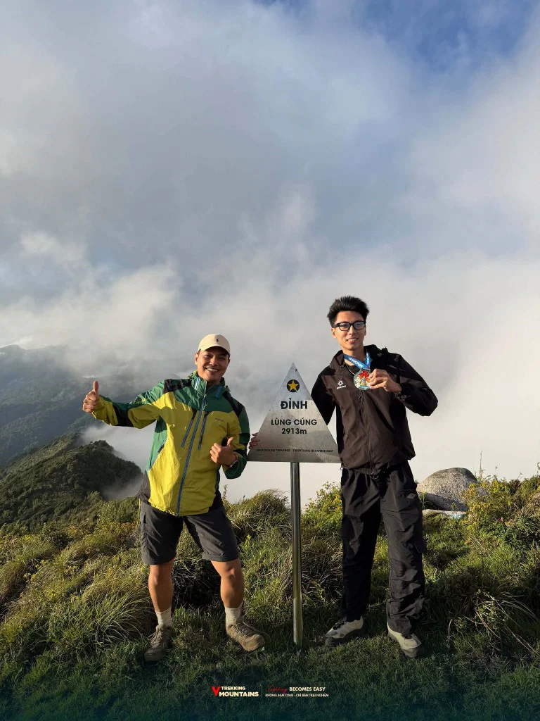 Hai trekker giơ ngón cái và huy chương bên cột mốc đỉnh Lùng Cúng 2913m, phía sau là mây phủ núi