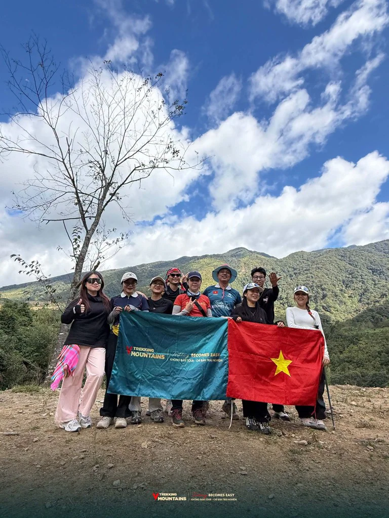 Đoàn trekking VMountains cầm cờ Việt Nam trước khi leo Lùng Cúng, phía sau là núi rừng và trời xanh