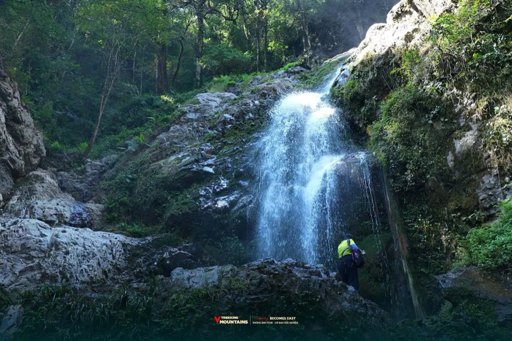 Trekker áo vàng đứng dưới thác nước giữa rừng trên cung trekking Lùng Cúng