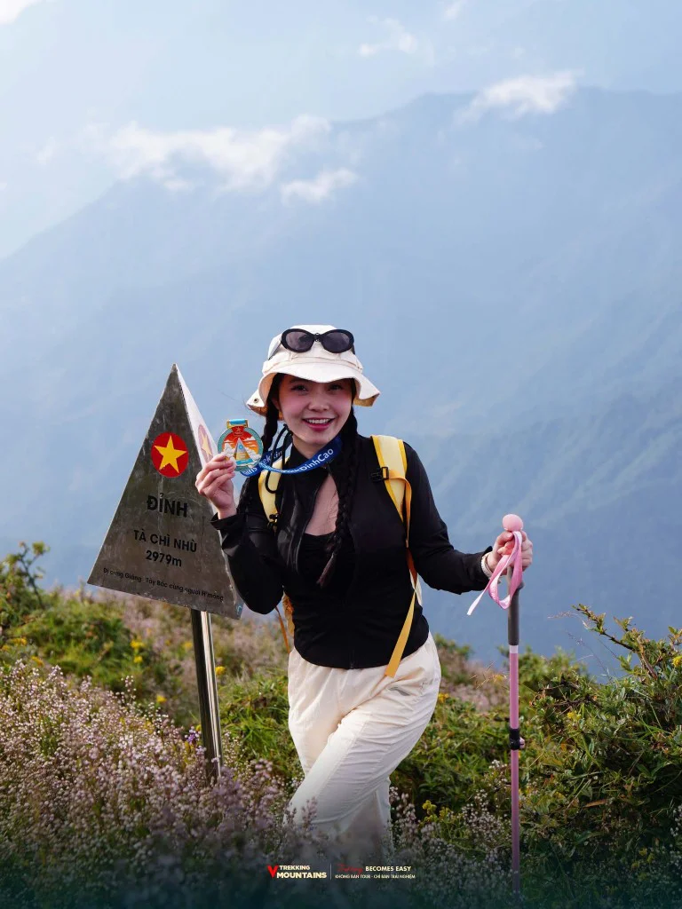 Nữ trekker cầm huy chương bên cột mốc đỉnh Tà Chì Nhù 2979m giữa thảm hoa Chi Pâu tím tháng 10